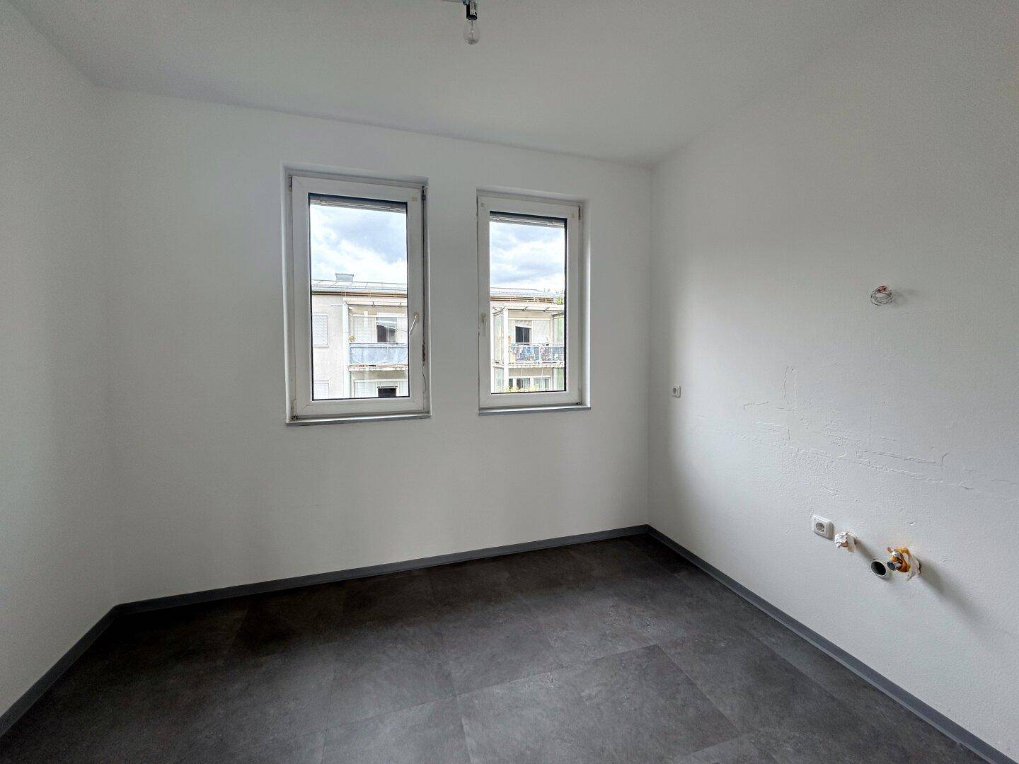 Heller Raum mit dunklem Fliesenboden und zwei Fenstern, vorbereitet für die Installation einer Küche.