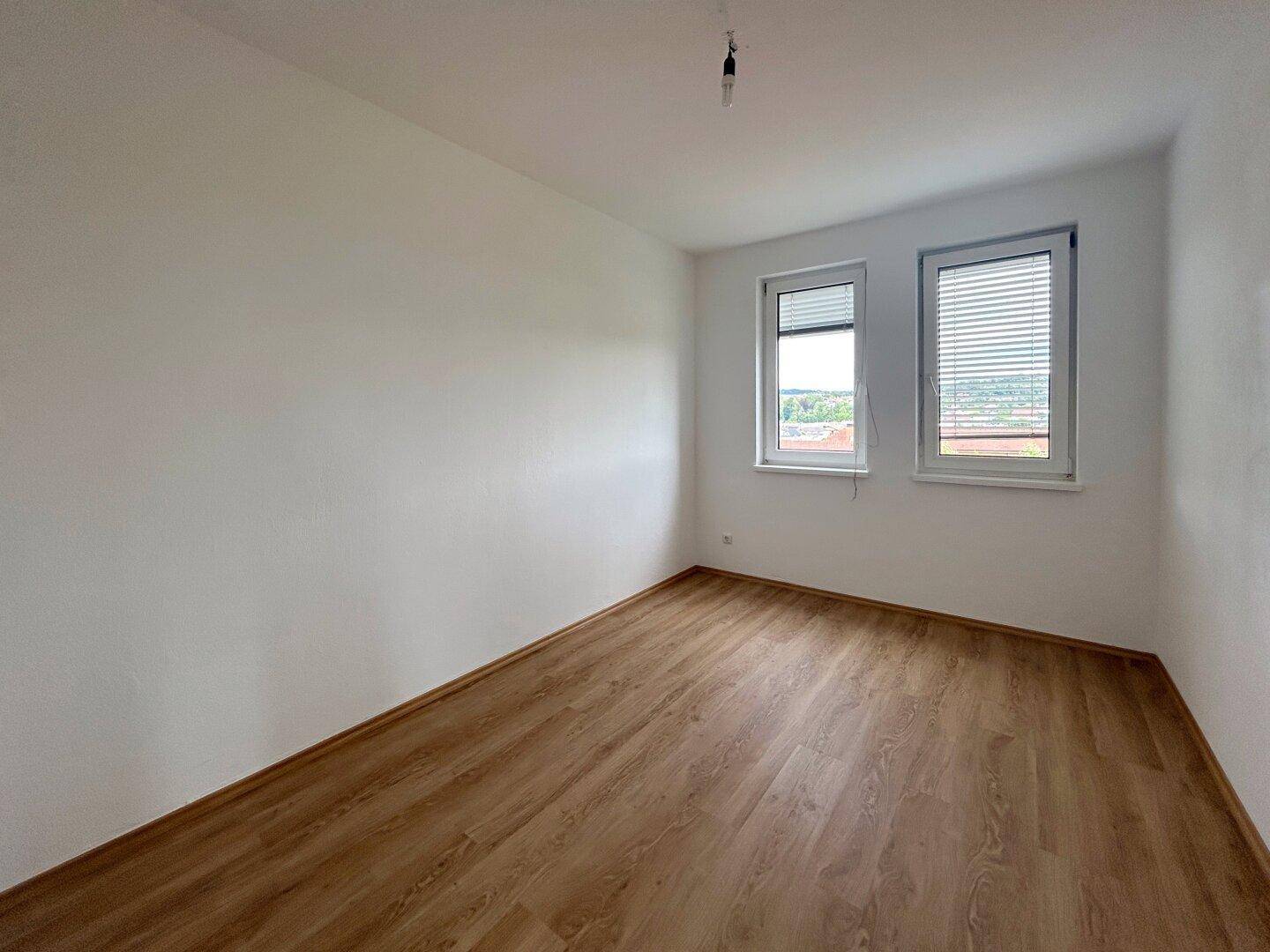 Helles Schlafzimmer mit Laminatboden und zwei Fenstern, die für ausreichend Tageslicht sorgen.