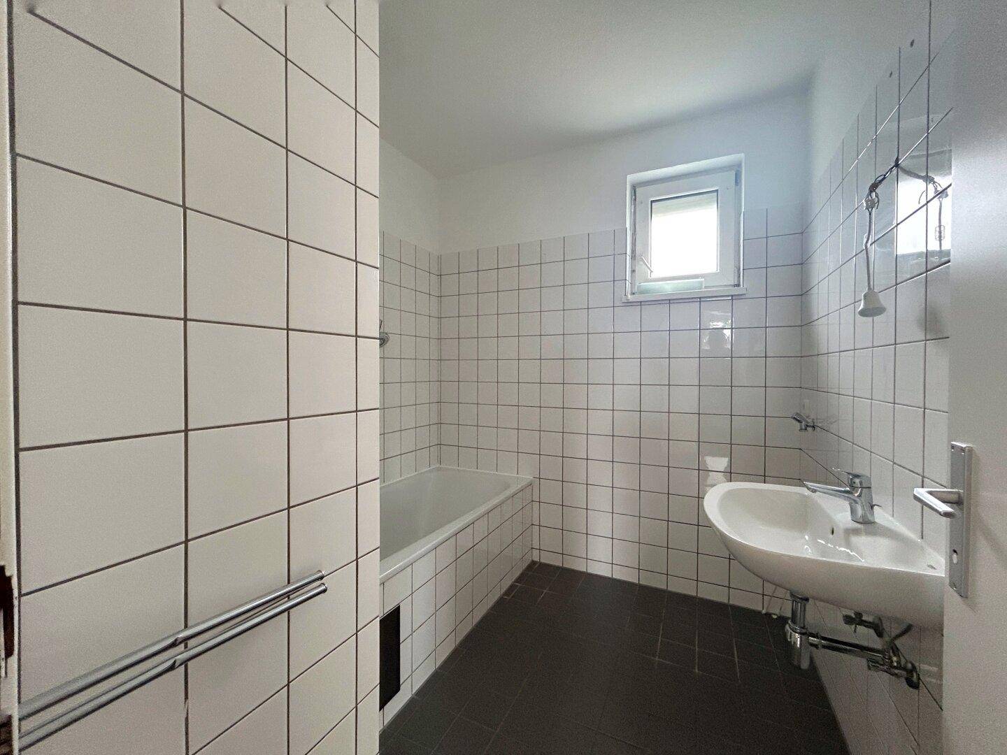 Badezimmer mit weißen Wandfliesen, dunklem Boden, Badewanne, Waschbecken und einem Fenster für Tageslicht.