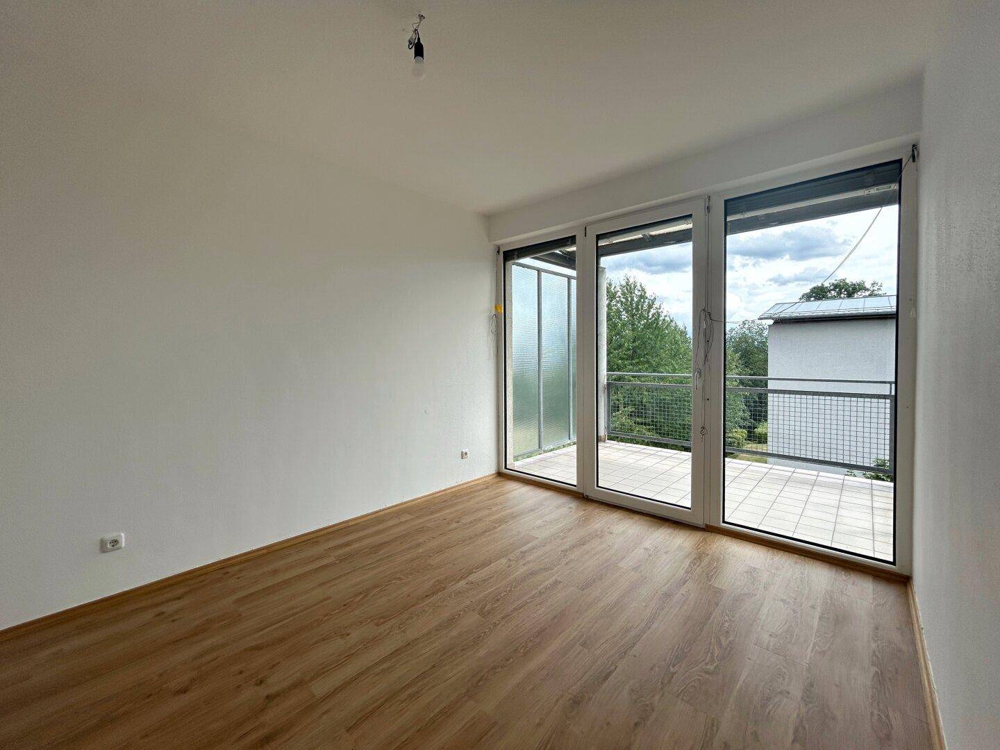 Heller, leerer Raum mit Laminatboden und großen Glasfronten, die zu einem Balkon oder einer Terrasse führen.