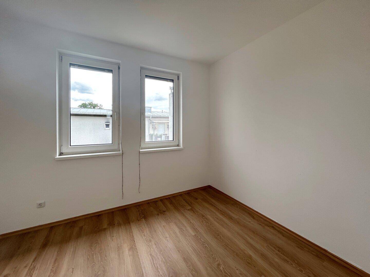 Leeres Zimmer mit Laminatboden und zwei Fenstern, ideal als Schlaf- oder Arbeitszimmer nutzbar.