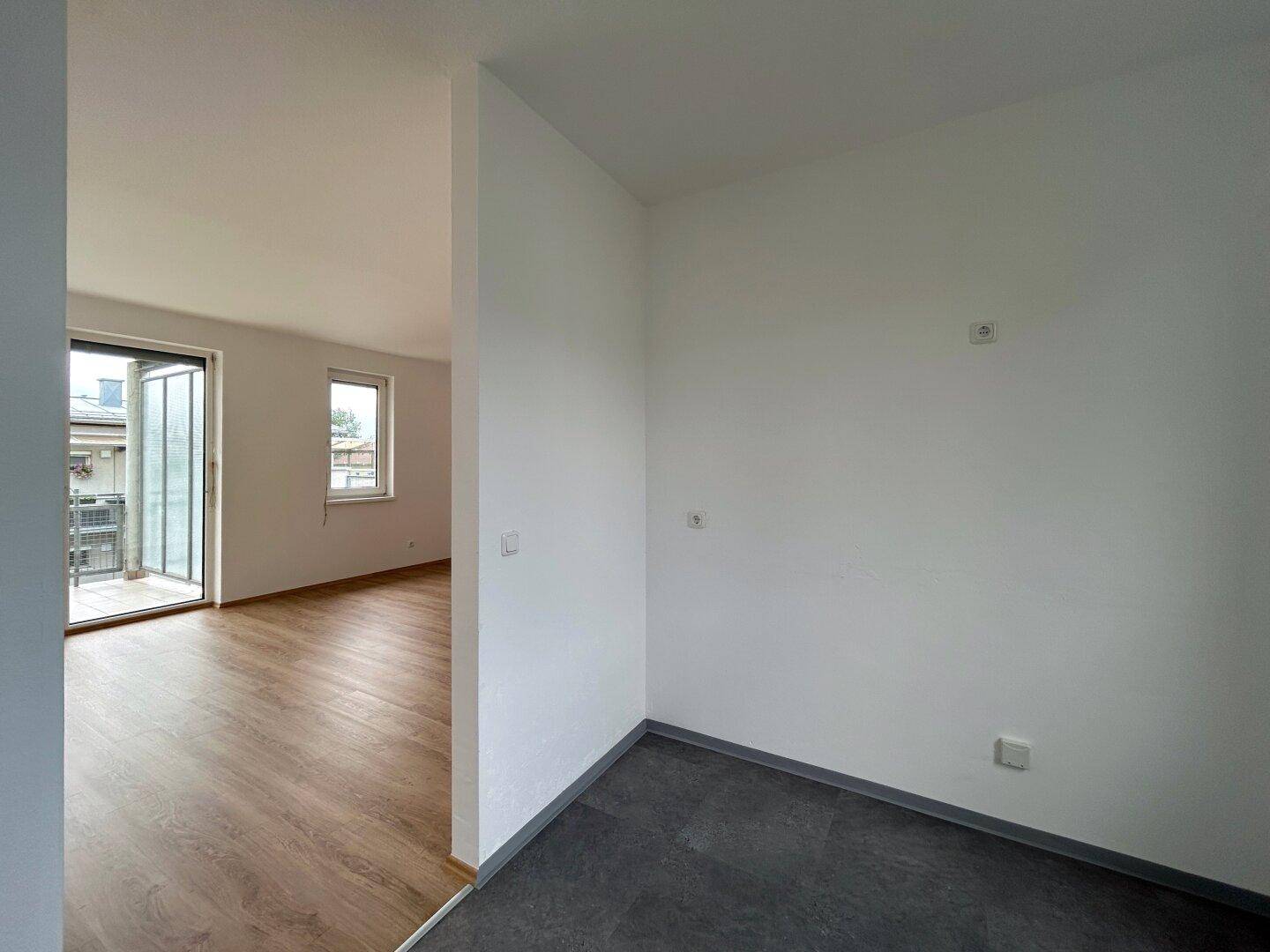 Unmöblierter Raum mit dunklem Fliesenboden und Küchenanschlüssen, angrenzend an einen hellen Wohnbereich.