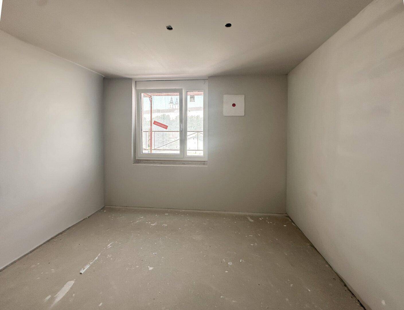 Unfertiges Zimmer mit Fenster, weißen Wänden und Betonboden.