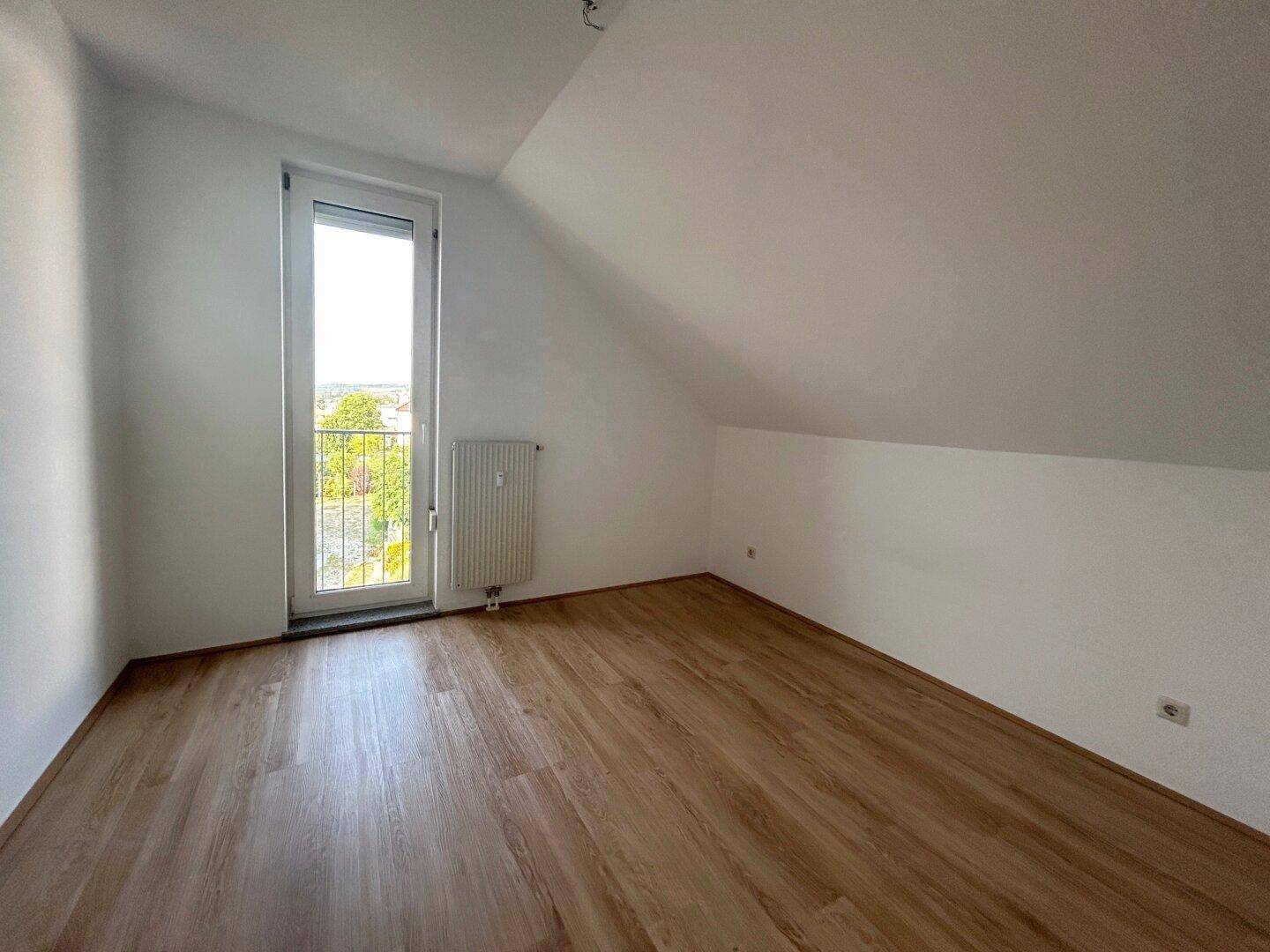 Helles Schlafzimmer mit Dachschräge, Laminatboden und Zugang zu einem französischen Balkon.