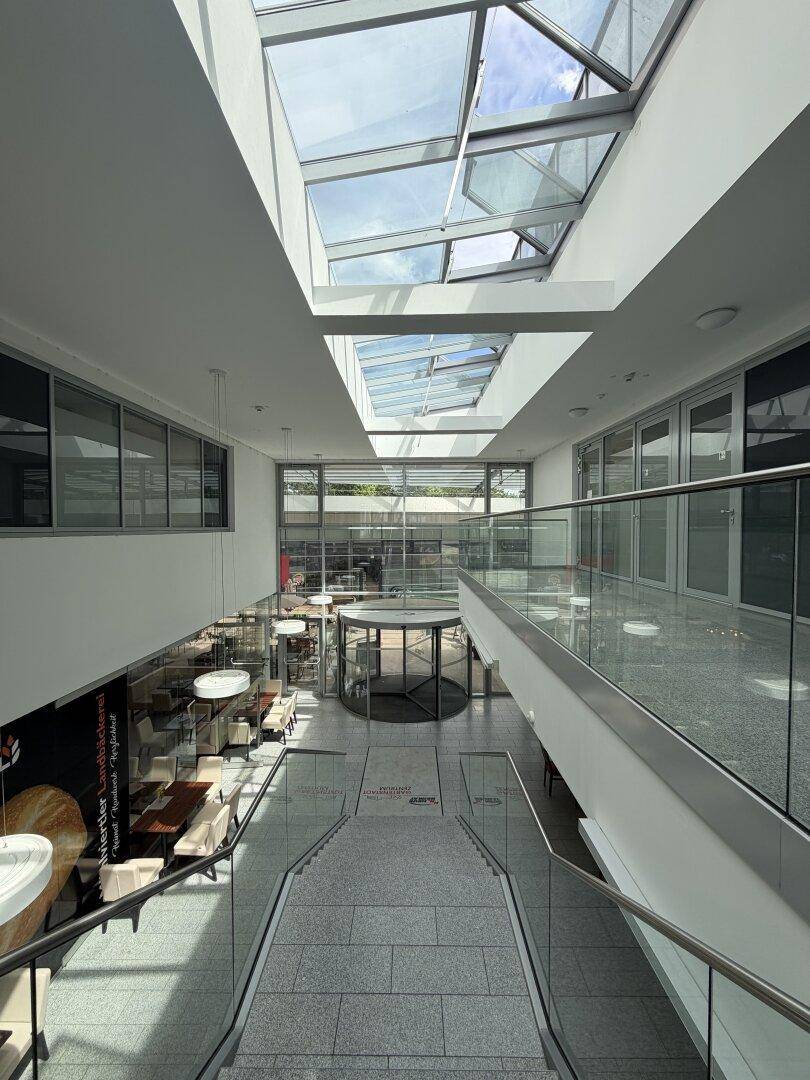 Großzügiges Atrium eines Geschäftsgebäudes mit Glasdach, Treppen und Blick auf die unteren Etagen.