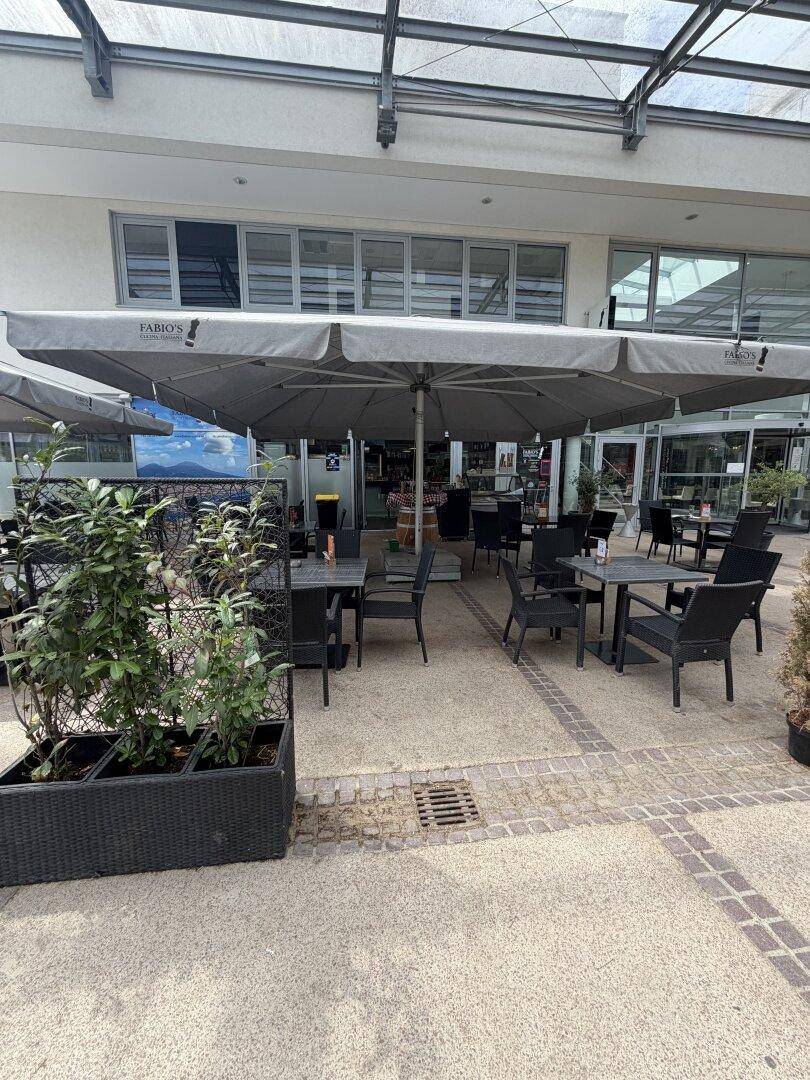 Detailansicht der Außenterrasse eines Cafés mit Tischen, Stühlen und großen Sonnenschirmen.