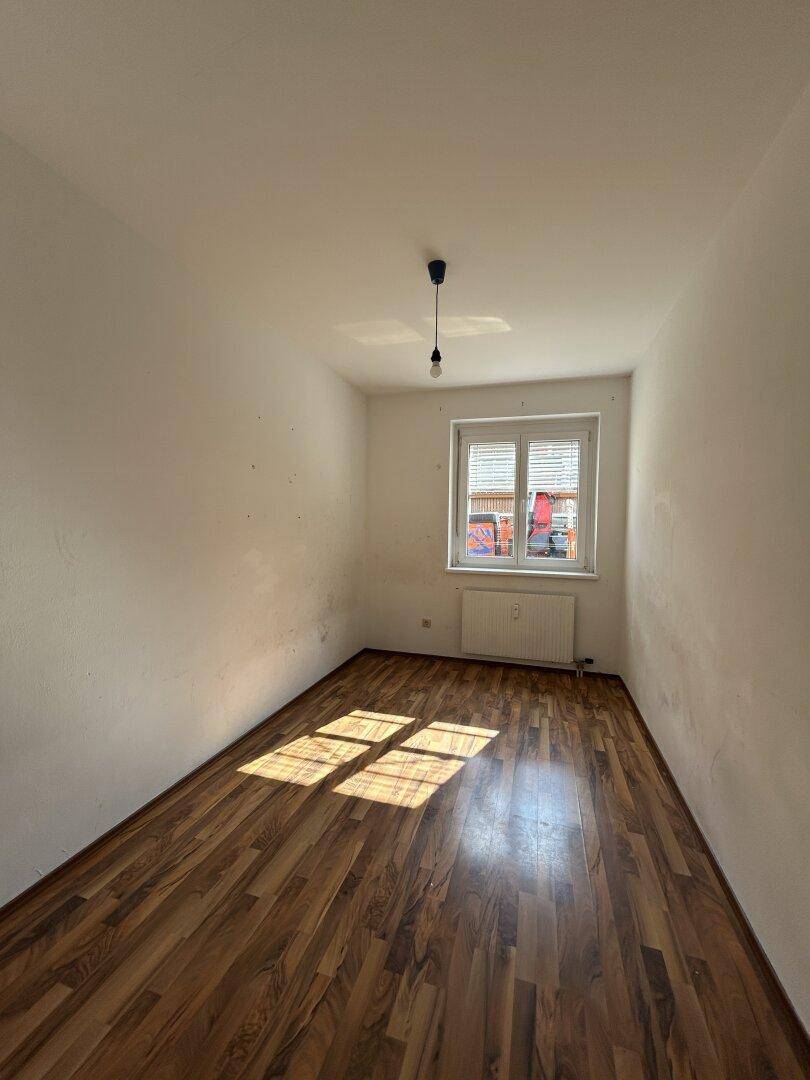 Leeres Zimmer mit Laminatboden, weiß gestrichenen Wänden und Fenster mit Jalousien, durch das Sonnenlicht fällt.