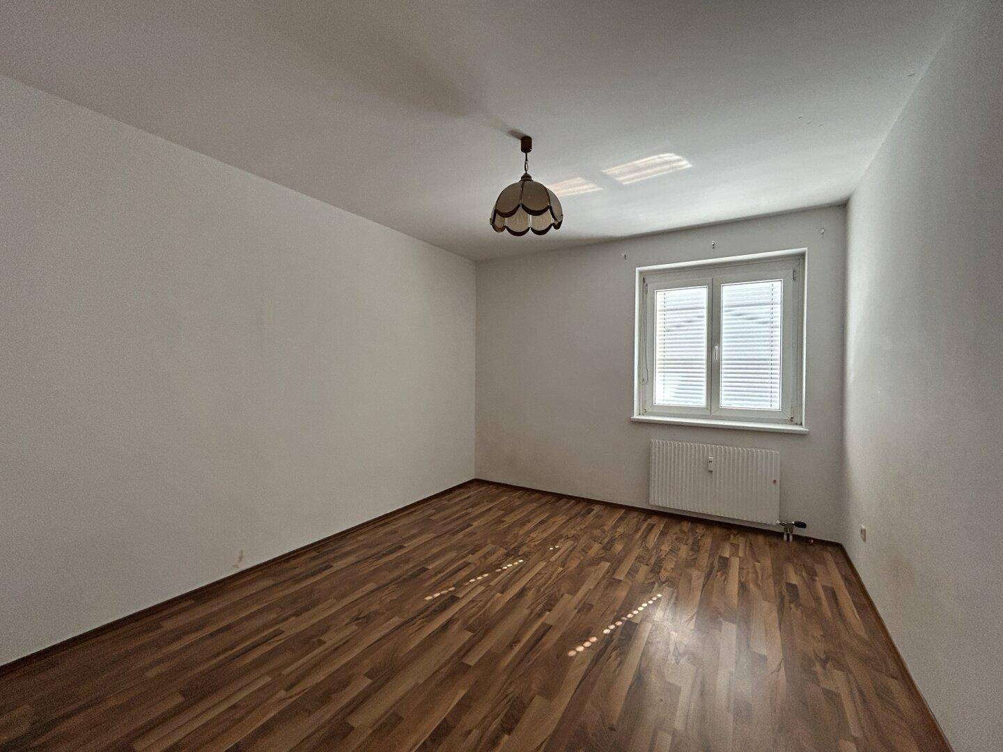 Helles, leeres Zimmer mit Laminatboden, weiß gestrichenen Wänden und einem Fenster mit Jalousien.