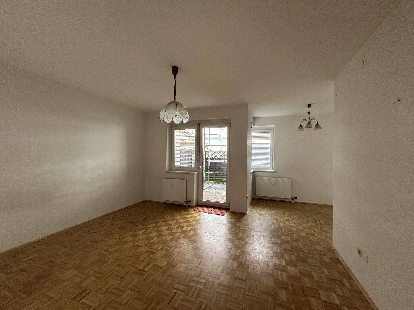 Geräumiges Wohnzimmer mit Parkettboden, weiß gestrichenen Wänden und Zugang zum Außenbereich durch eine Tür.
