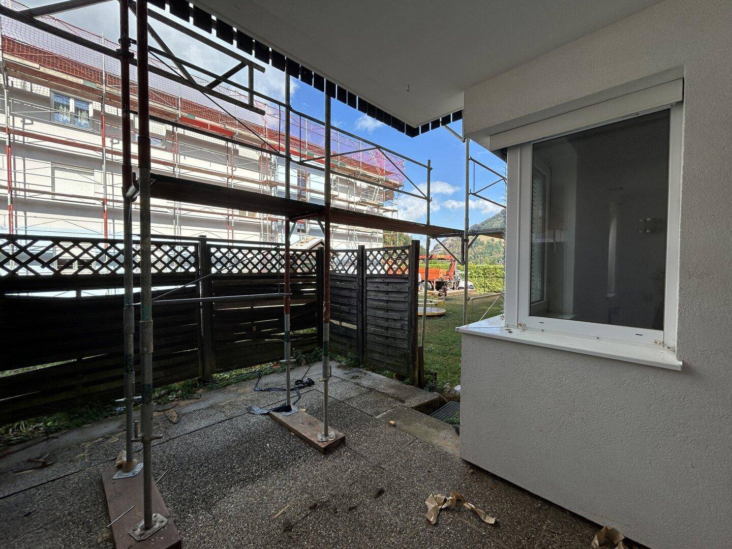Außenbereich mit Betonboden, Holzzaun und Fenster mit Rollladen, umgeben von Baugerüsten.