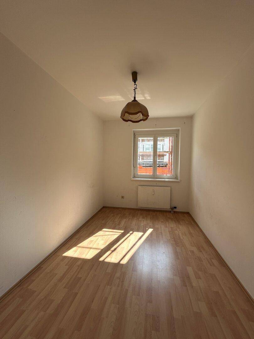 Leeres Schlafzimmer mit Laminatboden, weiß gestrichenen Wänden und einem Fenster mit viel Tageslicht.