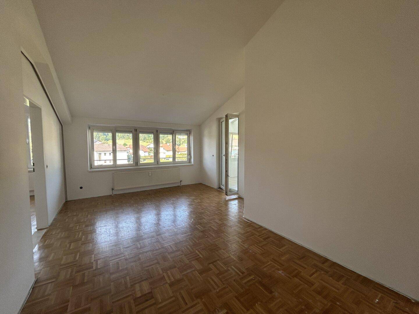 Geräumiges, leeres Wohnzimmer mit Parkettboden, weißen Wänden und Zugang zu einem Außenbereich.