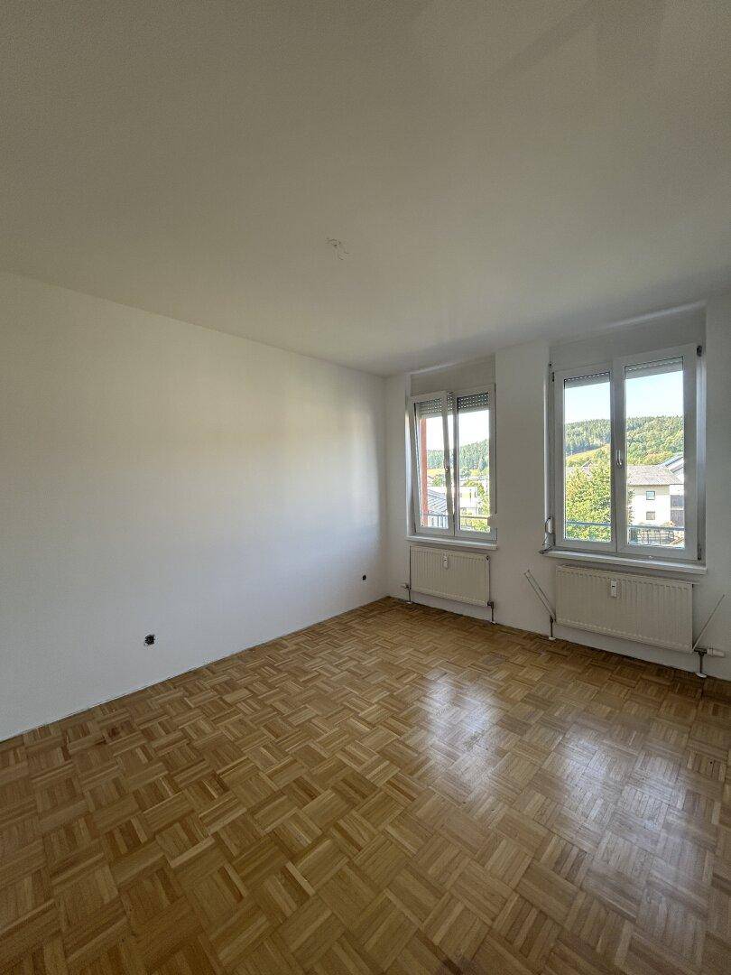 Helles, leeres Schlafzimmer mit Parkettboden und zwei Fenstern, bereit zur individuellen Gestaltung.