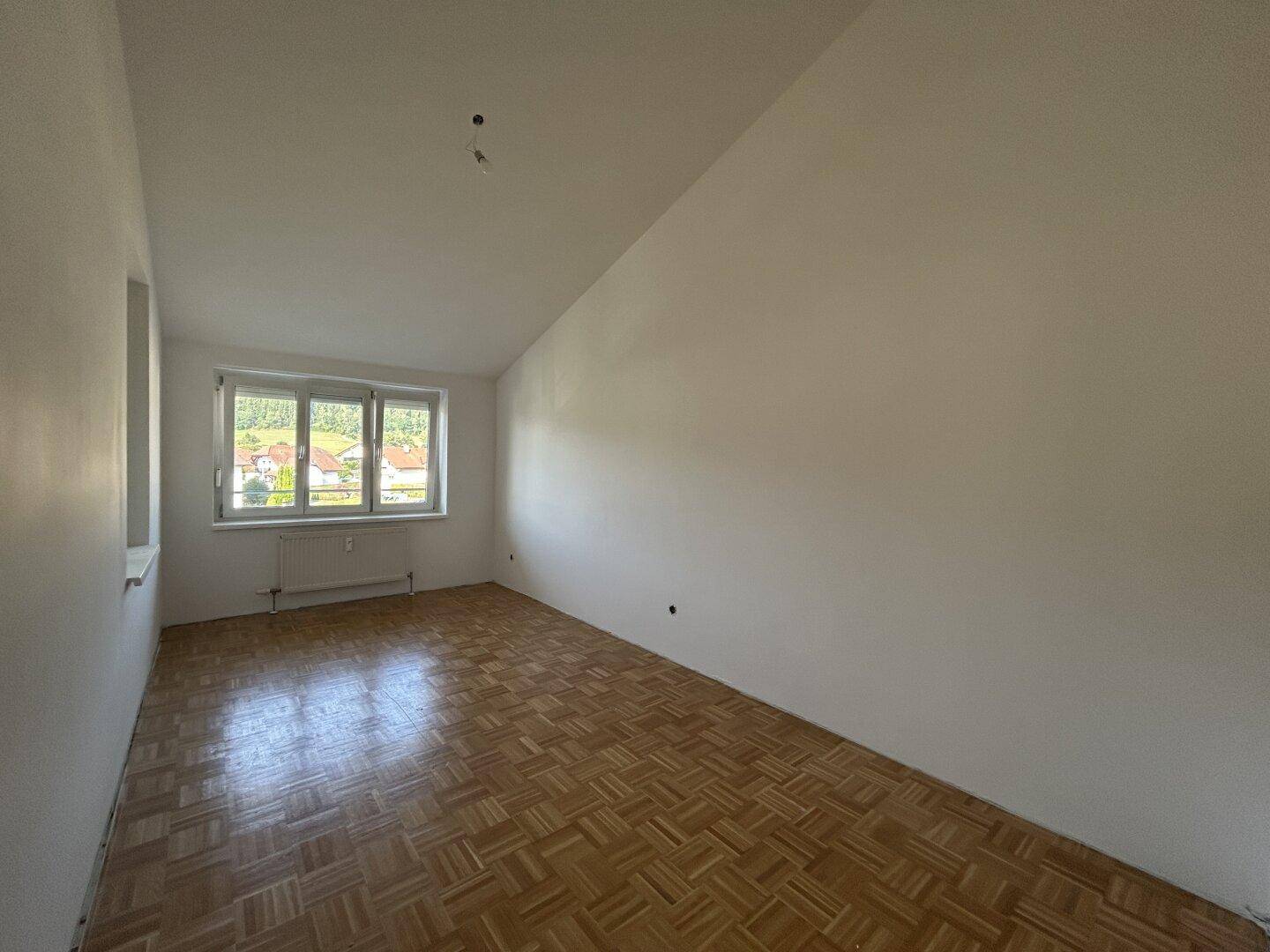 Geräumiges, leeres Zimmer mit Parkettboden, weißen Wänden und einem Fenster unter der Dachschräge.