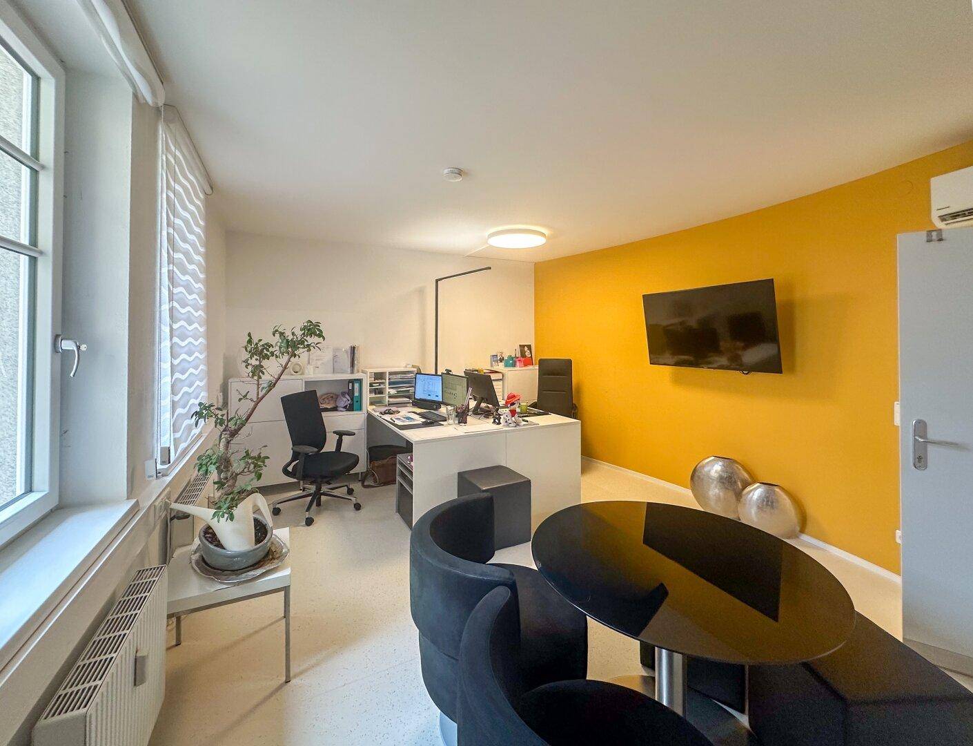 Modernes Büro mit gelber Akzentwand, Schreibtisch und kleinem Besprechungstisch.