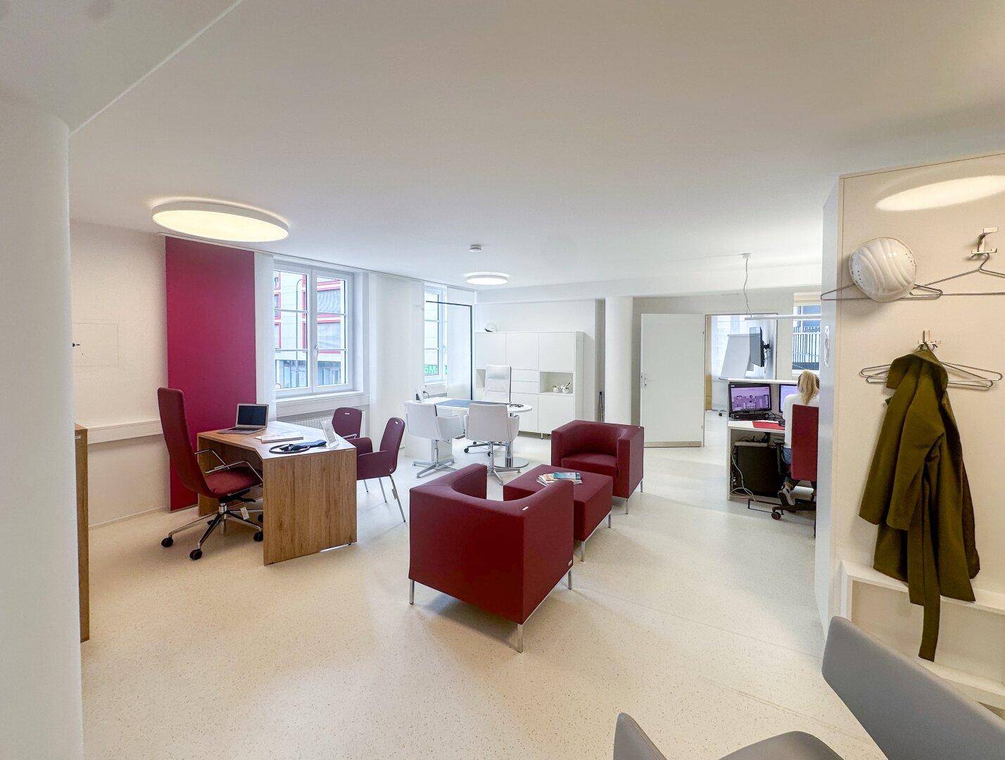 Helles und geräumiges Büro mit verschiedenen Arbeitsbereichen, modernen Möbeln und roten Akzenten.