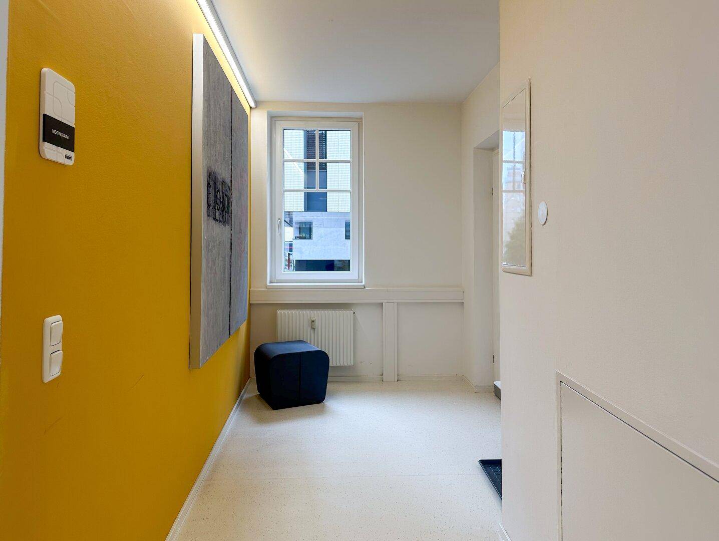 Heller Flur mit gelber Akzentwand, Fenster und einem kleinen Sitzhocker.