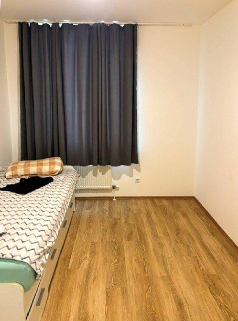 Schlafzimmer mit Laminatboden, einem Bett und einem Fenster mit dunklen Vorhängen.