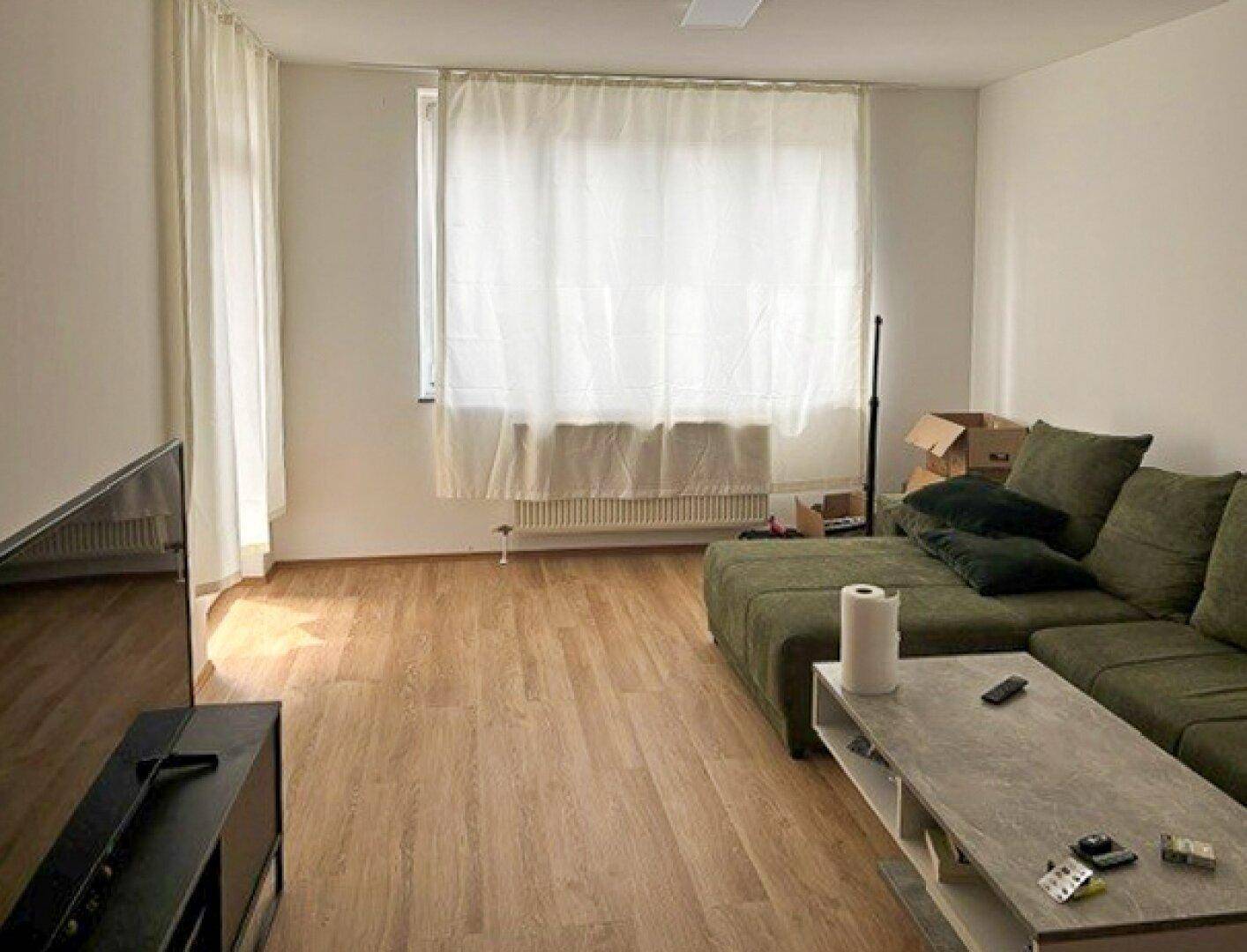 Helles Wohnzimmer mit Laminatboden, einem Sofa und einem Fenster mit hellen Vorhängen.