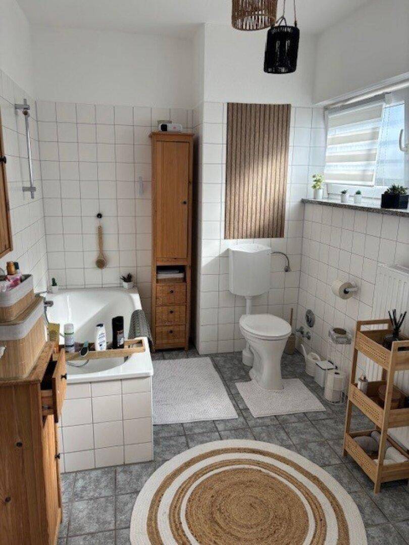 Helles Badezimmer mit weißen Fliesen, Badewanne, Toilette und Holzmöbeln.