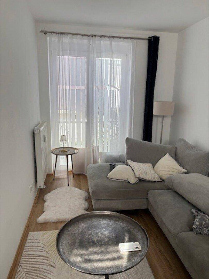 Helles Wohnzimmer mit grauem Ecksofa, modernen Beistelltischen und Fenster mit Vorhängen.