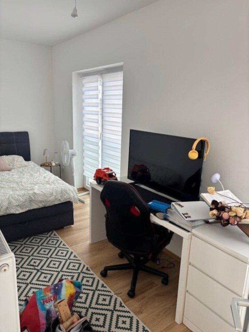 Schlafzimmer mit Bett, Schreibtisch, Computer und einem großen Fenster.
