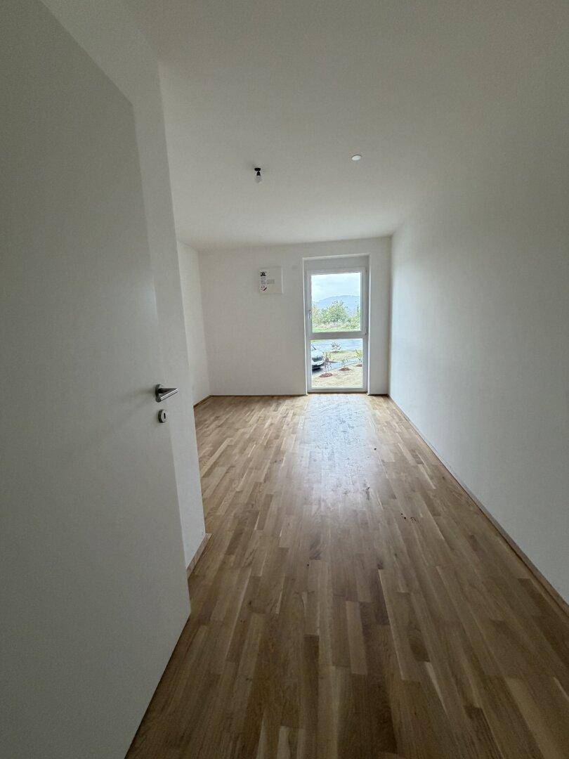 Leerer Raum mit Holzboden und einem großen Fenster, das viel Licht hereinlässt. Ideal als Schlaf- oder Arbeitszimmer.