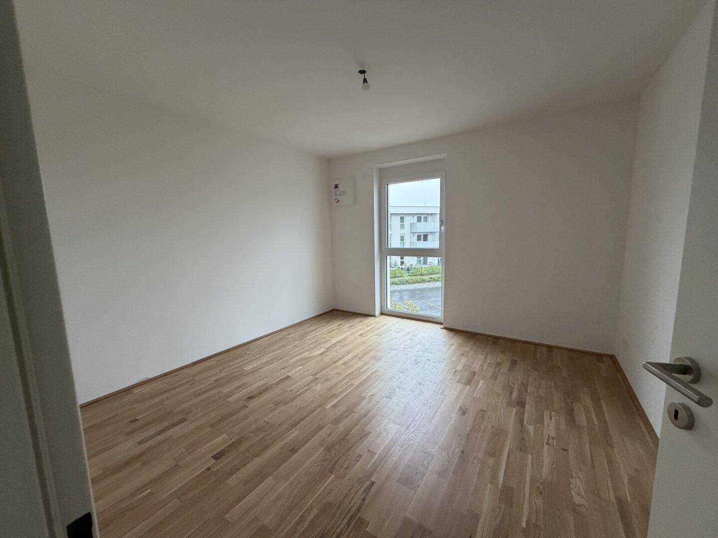 Leeres Schlafzimmer mit hellem Parkettboden und einem großen Fenster, das viel Tageslicht hereinlässt.