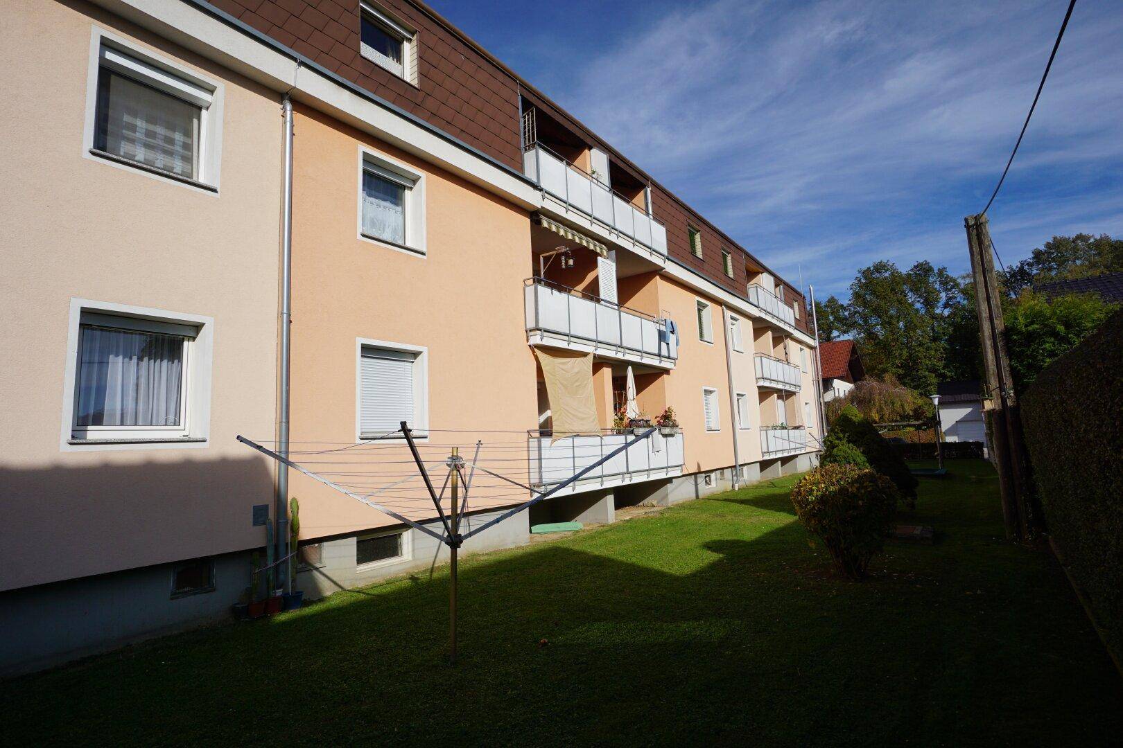 Seitenansicht eines Mehrfamilienhauses mit Balkonen, Wäscheleine und einer gepflegten Rasenfläche unter blauem Himmel.