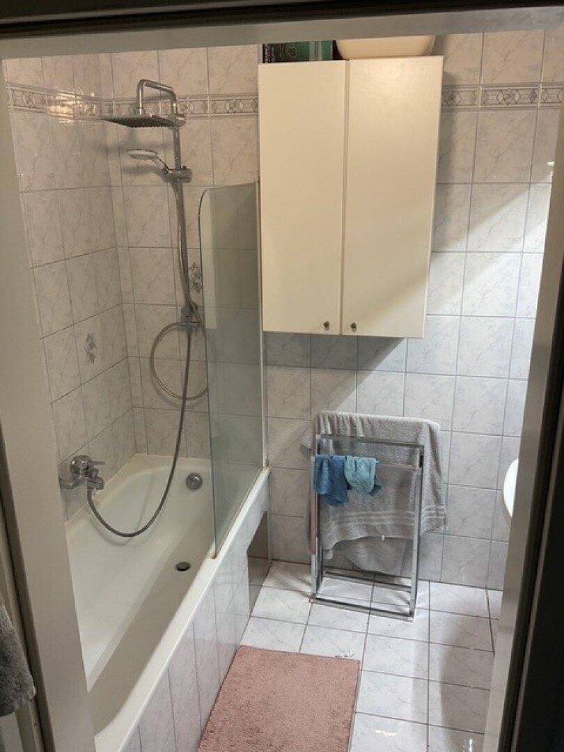 Badezimmer mit Badewanne, Duschvorrichtung, hellen Fliesen und einem praktischen Wandschrank für Stauraum.