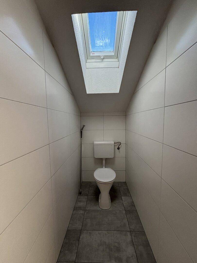 Kleines WC mit weißen Wandfliesen, grauen Bodenfliesen und einem Dachfenster.