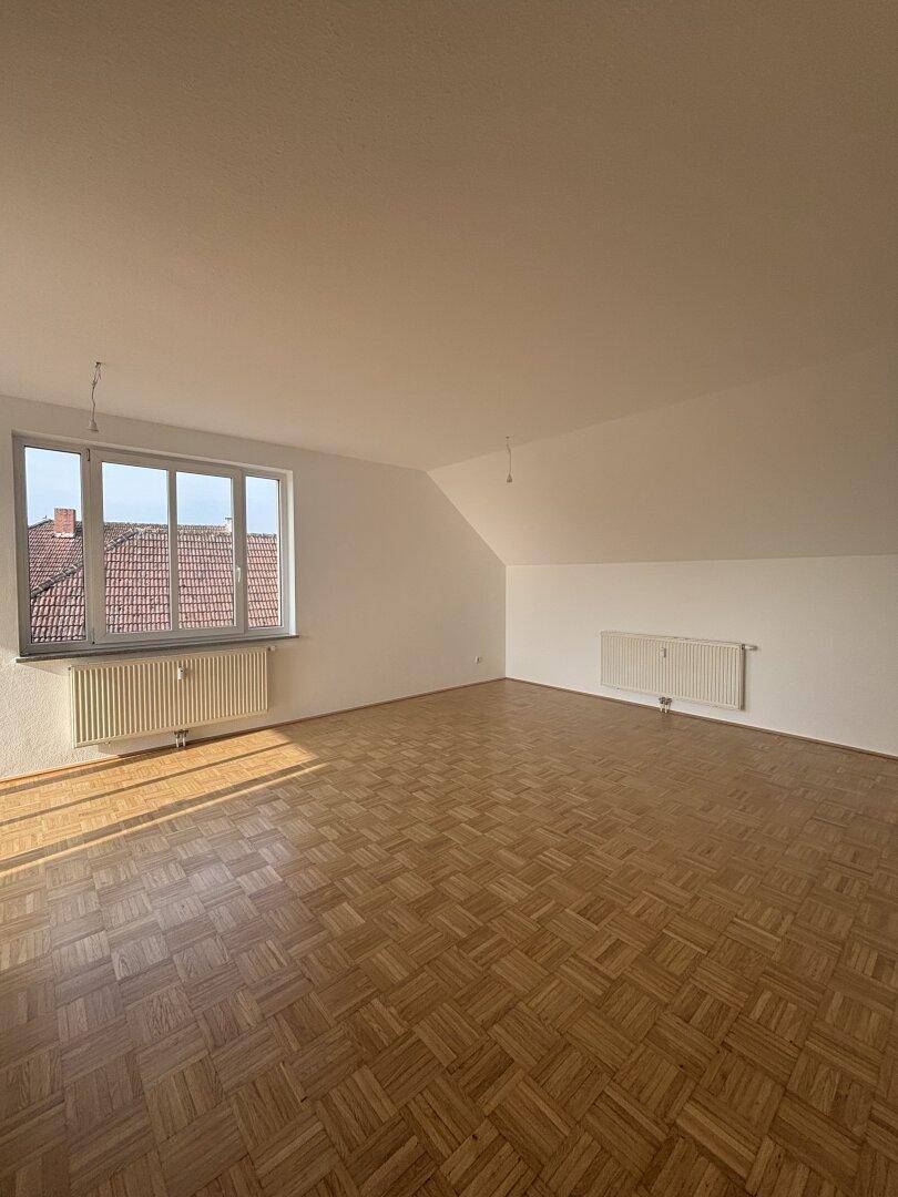 Heller, leerer Raum im Dachgeschoss mit Parkettboden, schrägen Wänden und großem Fenster.