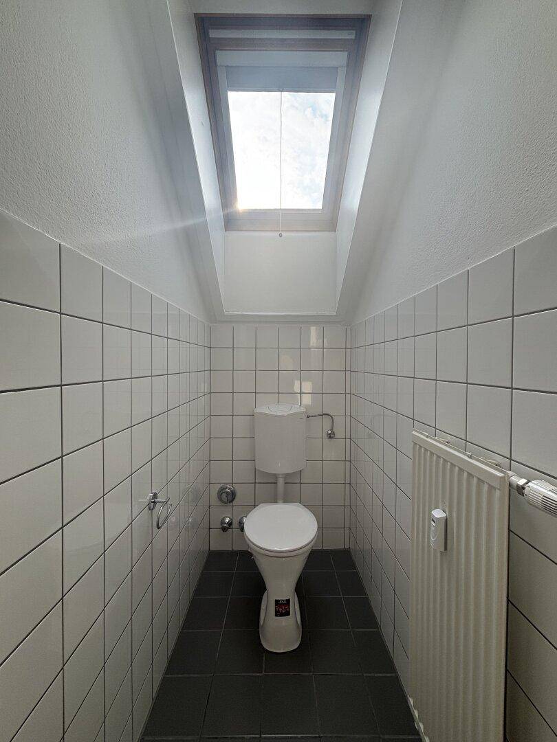 Kleines WC mit weißen Wandfliesen, dunklem Boden und einem Dachfenster für Tageslicht.