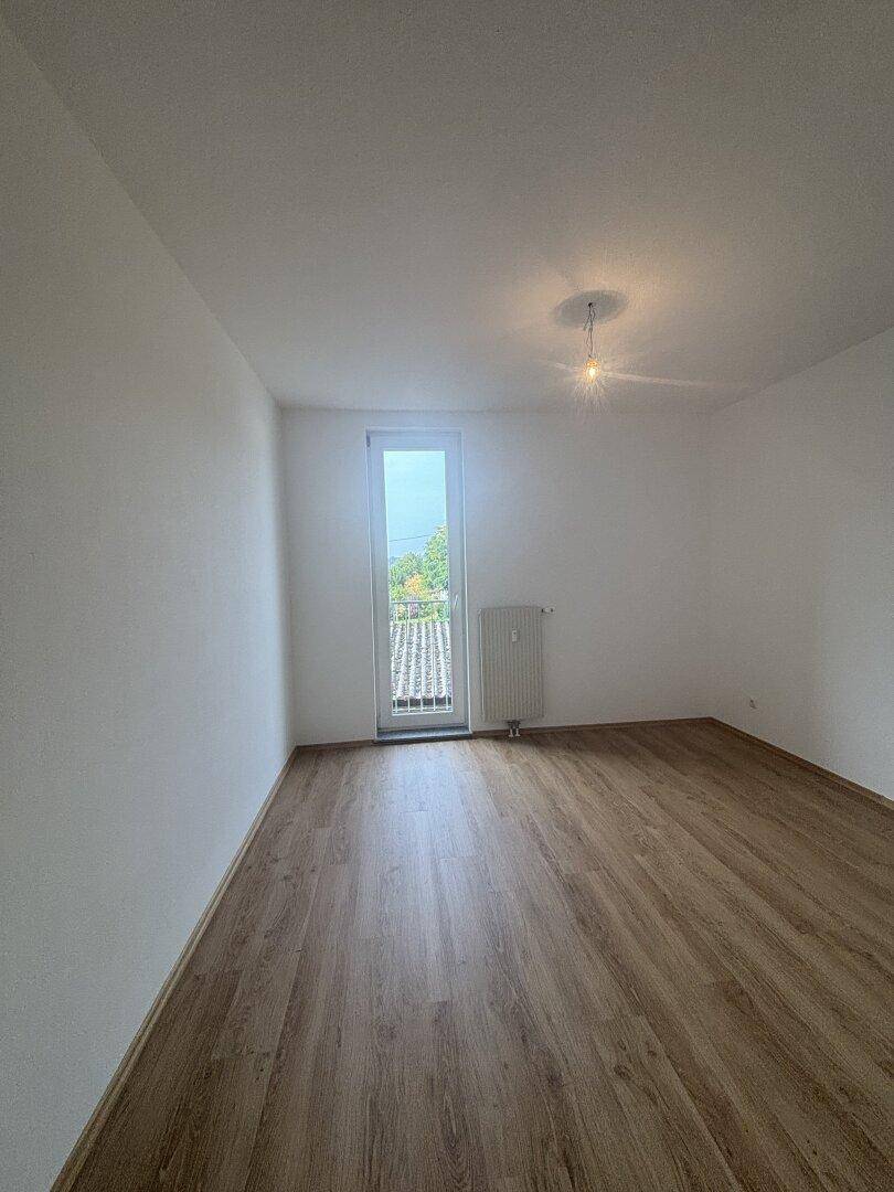 Leeres Zimmer mit Laminatboden und großem Fenster, geeignet als Schlaf- oder Arbeitszimmer.