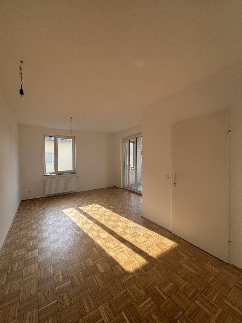 Lichtdurchflutetes, leeres Wohnzimmer mit Parkettboden und Zugang zum Balkon.