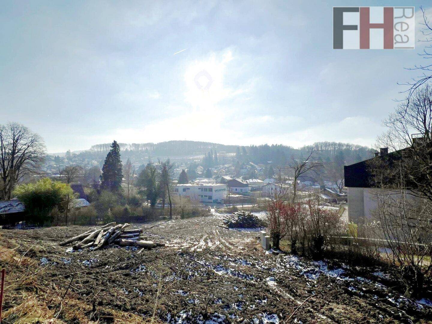 Weitläufiges Grundstück mit Holzstapeln und Blick auf die umliegenden Gebäude und bewaldeten Hügel an einem sonnigen Tag.