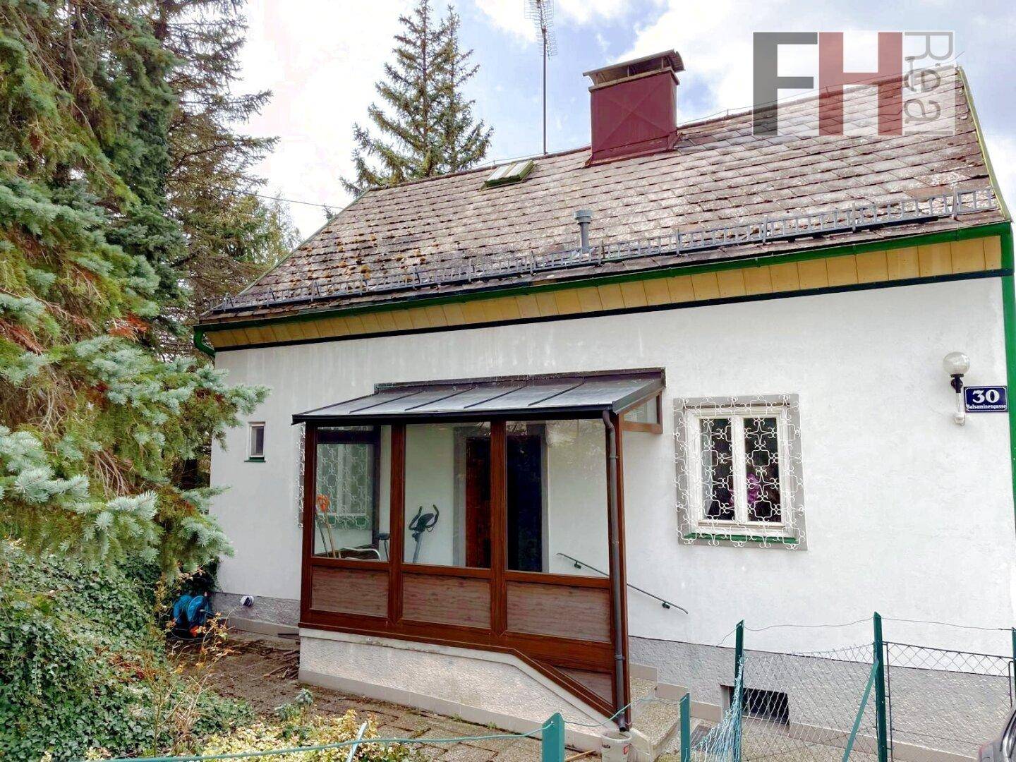Fassade eines Hauses mit einem kleinen, verglasten Eingangsbereich und Fensterläden an der Seite.