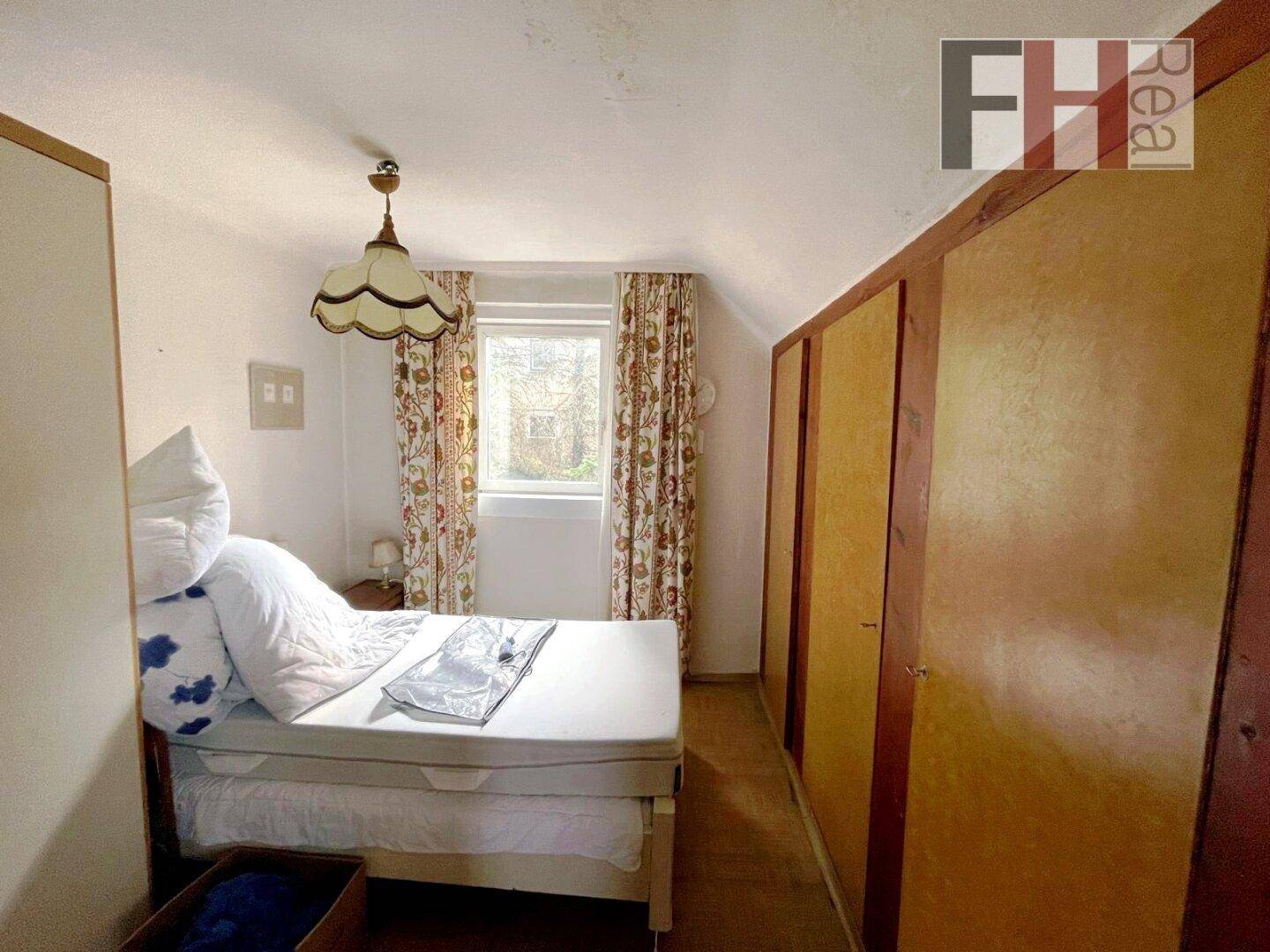 Schlafzimmer mit Dachschräge, Einbauschränken und einem Fenster mit gemusterten Vorhängen.