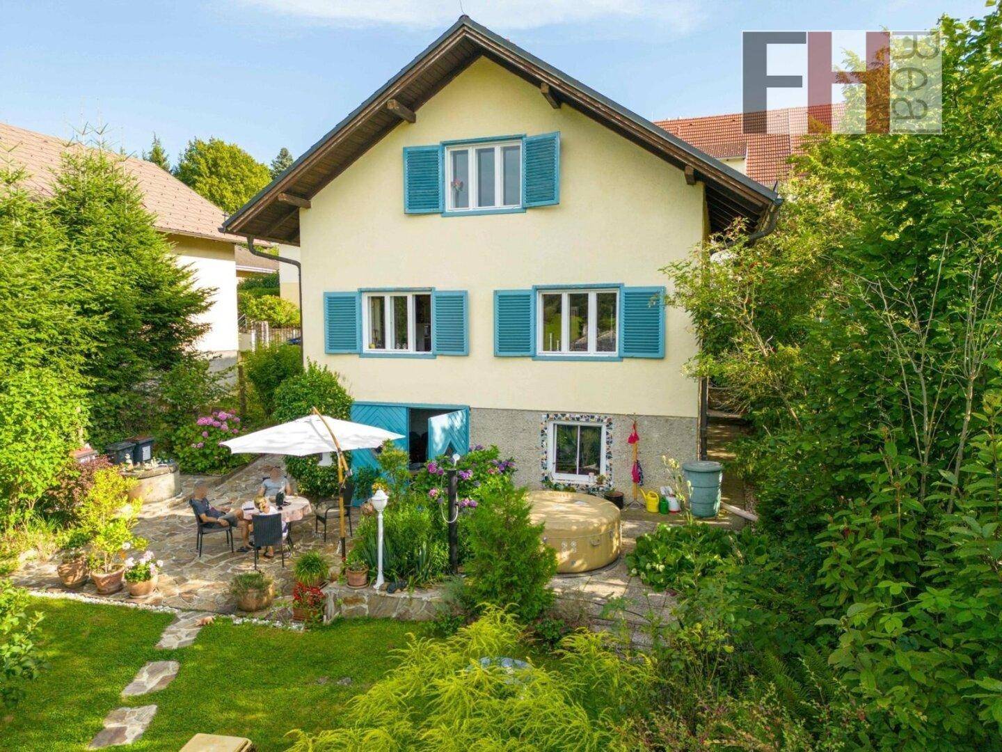Charmantes Haus mit blauen Fensterläden und einer gemütlichen Terrasse, umgeben von einem grünen Garten.