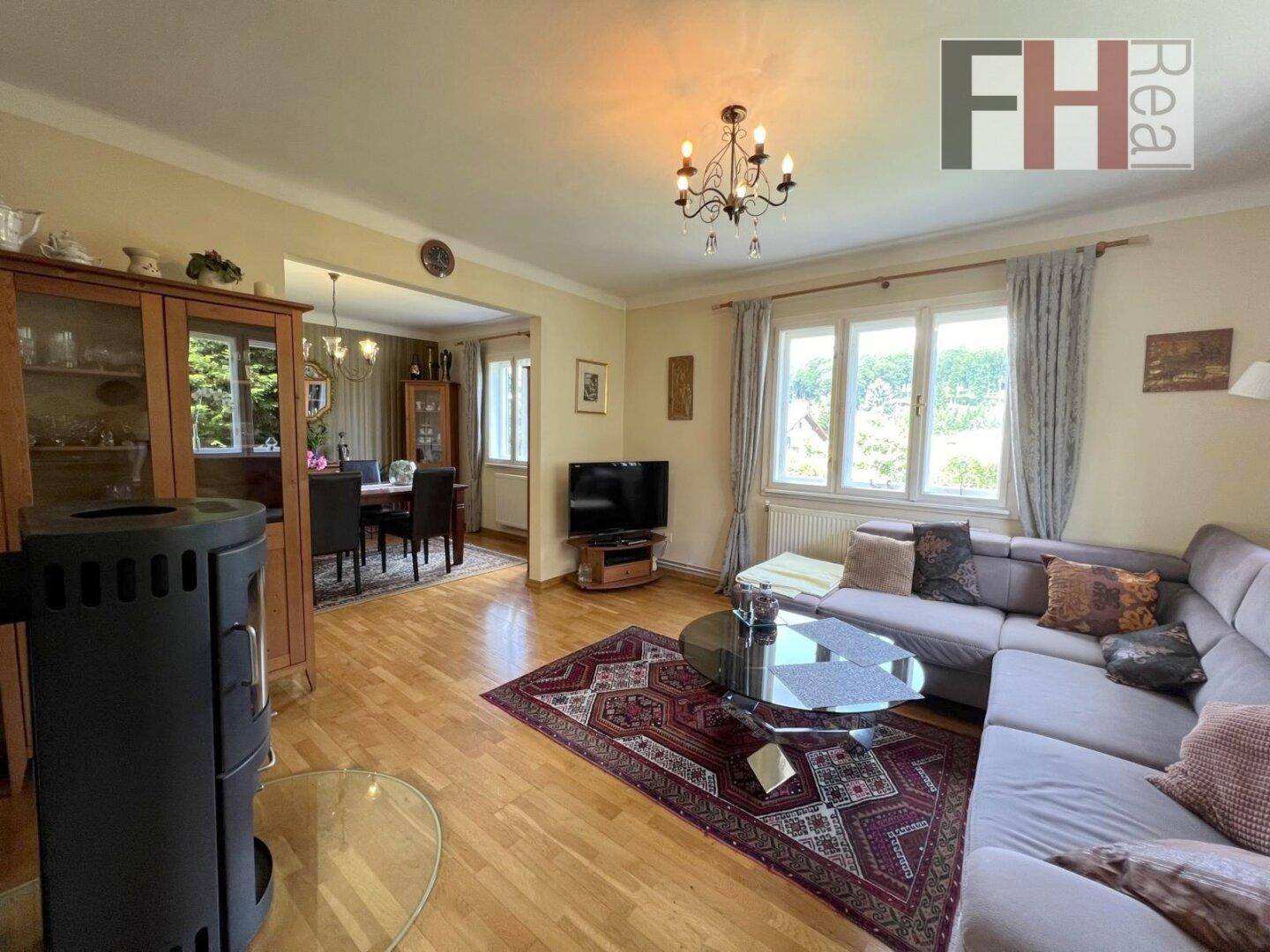 Helles Wohn- und Esszimmer mit Holzboden, Kaminofen und Blick ins Grüne durch große Fenster.