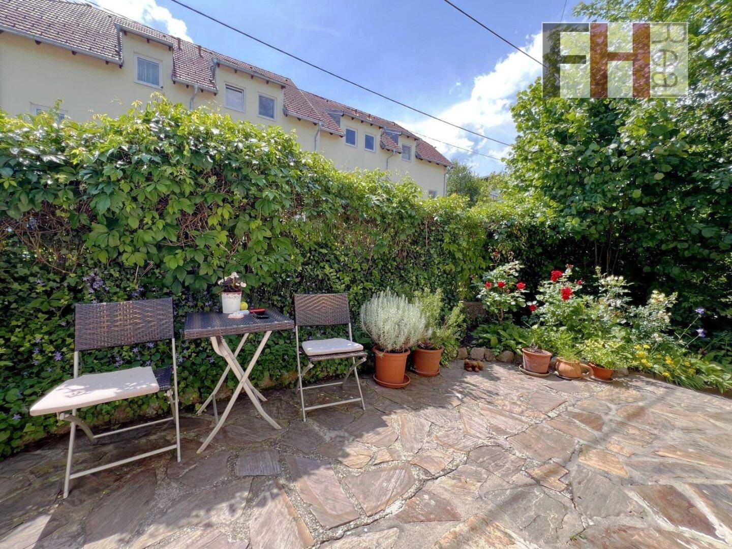 Gemütlicher gepflasterter Sitzbereich im Garten, umgeben von einer hohen Hecke und blühenden Rosen.