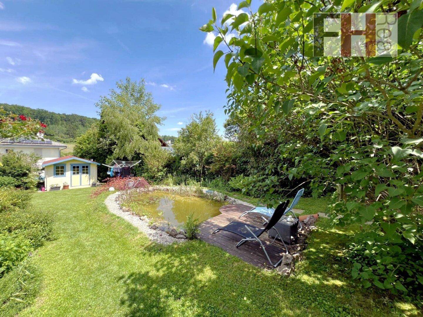 Idyllischer Gartenteich mit Holzdeck und Liegestühlen, umgeben von Rasen und üppiger Vegetation.