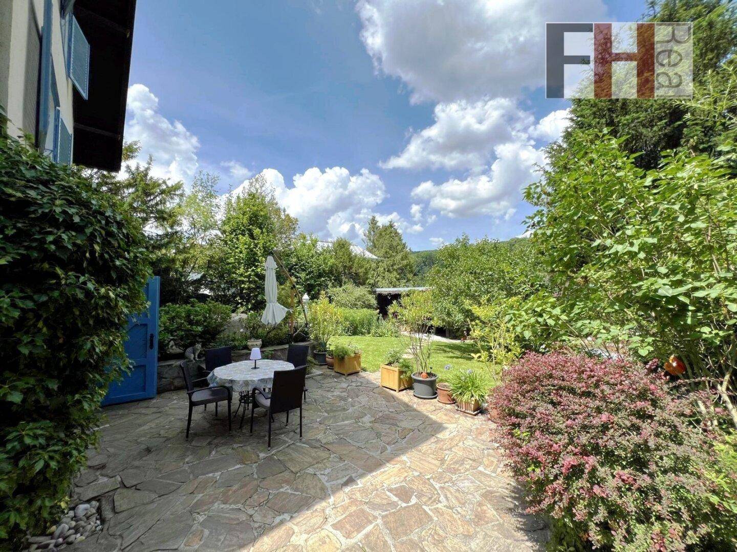 Große gepflasterte Terrasse mit Esstisch und Stühlen, umgeben von einem weitläufigen grünen Garten.