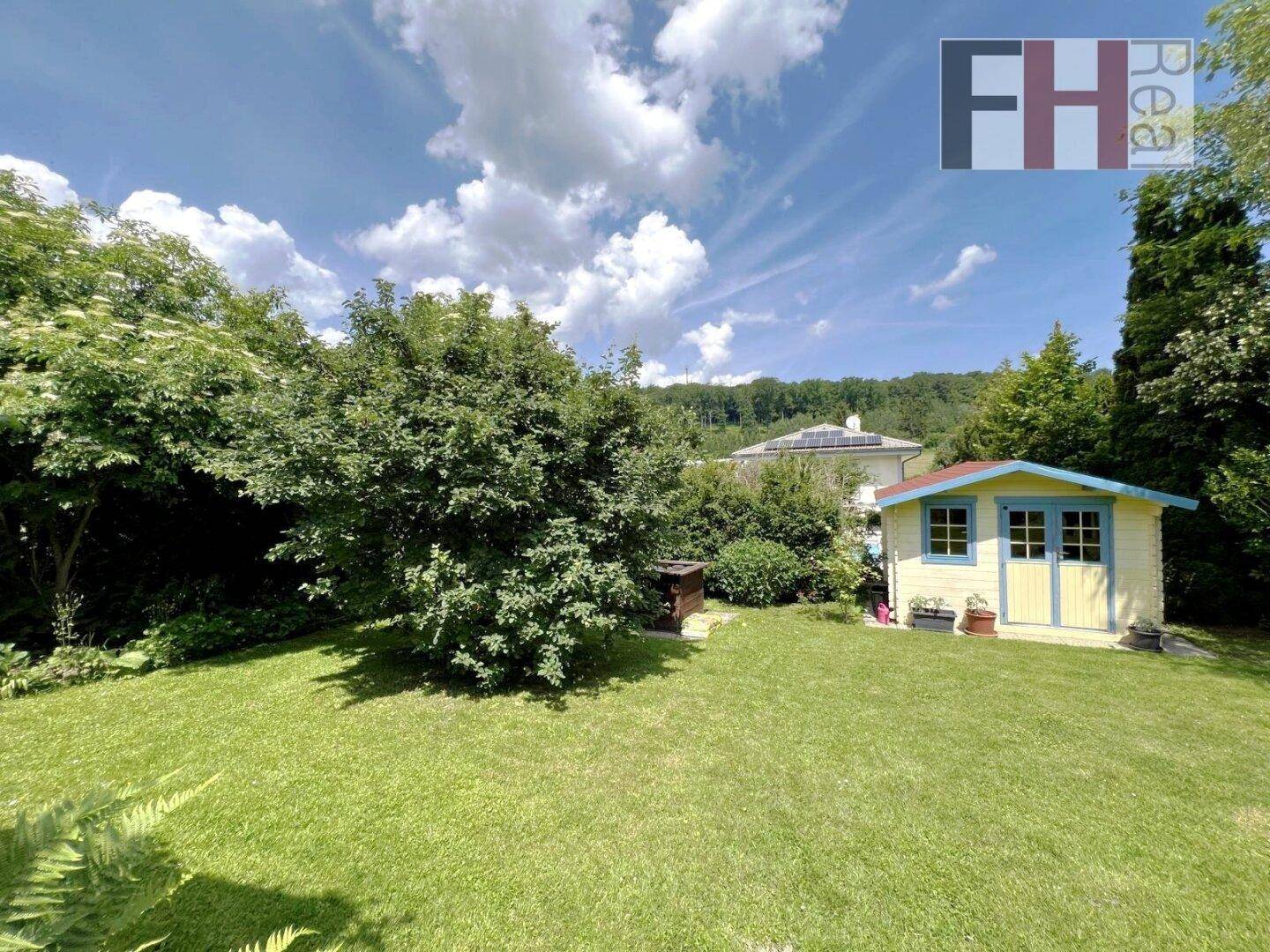 Großer, sonniger Garten mit Rasenfläche und einem charmanten gelben Gartenhaus, umgeben von Bäumen.