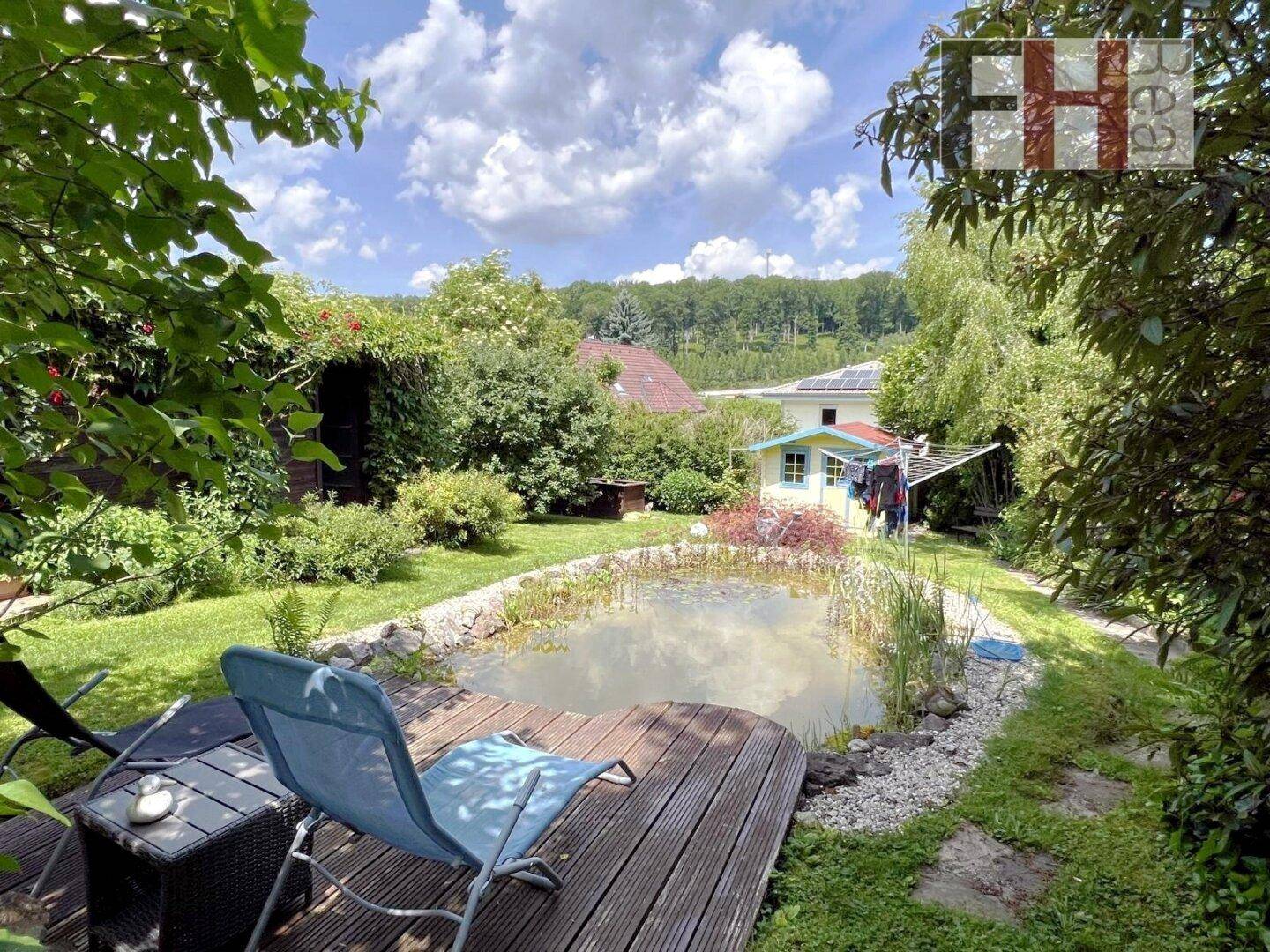 Entspannungsbereich am Gartenteich mit Holzdeck und Liegestühlen, umgeben von üppiger Natur.