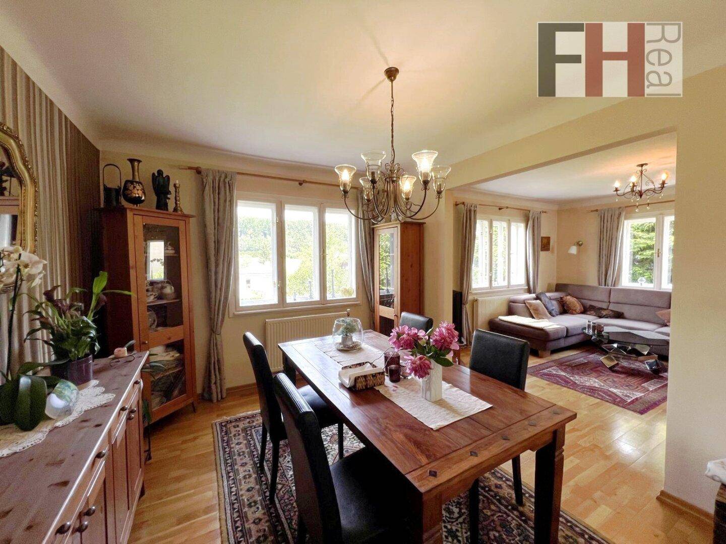 Geräumiger Essbereich mit Holztisch und Kronleuchter, offen zum Wohnzimmer mit Blick ins Grüne.