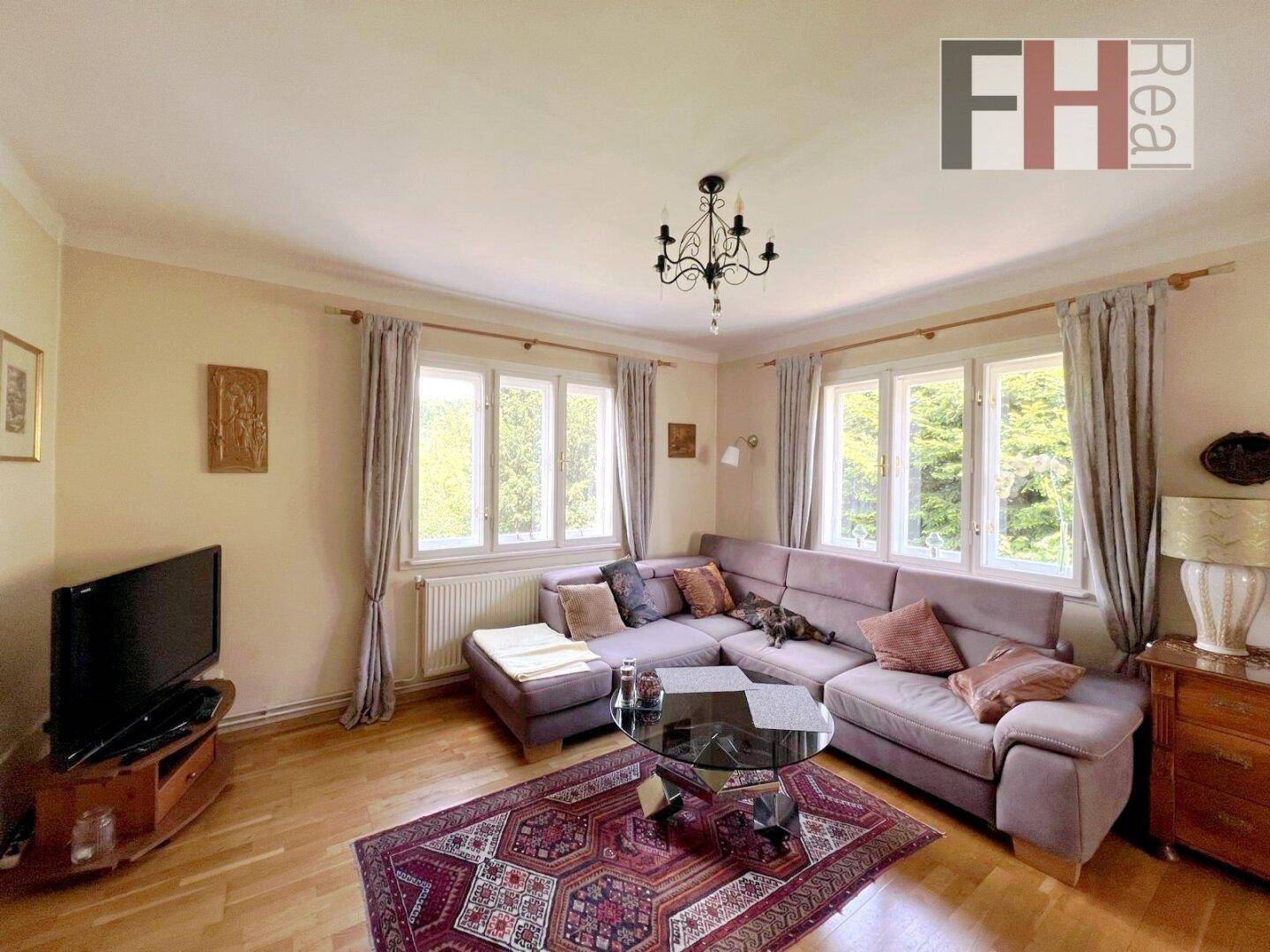 Helles Wohnzimmer mit gemütlichem Ecksofa, Holzboden und Blick ins Grüne durch mehrere Fenster.