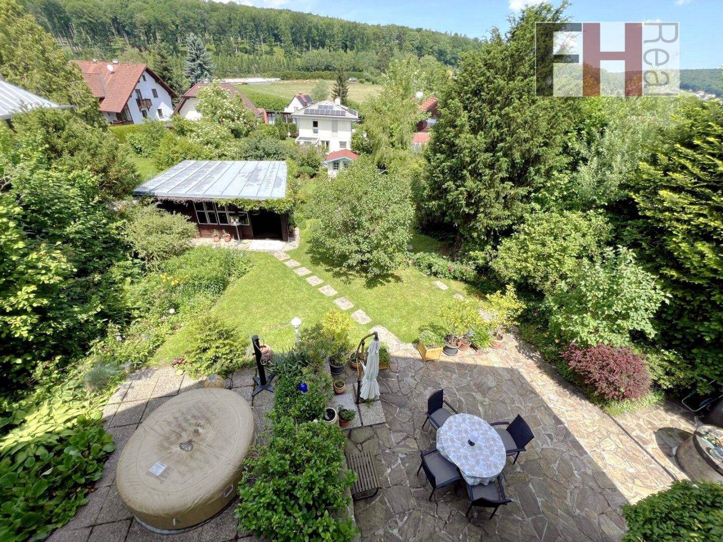 Große gepflasterte Terrasse mit Sitzbereich und Hot Tub, angrenzend an einen weitläufigen Garten mit Gartenhaus und grüner Umgebung.
