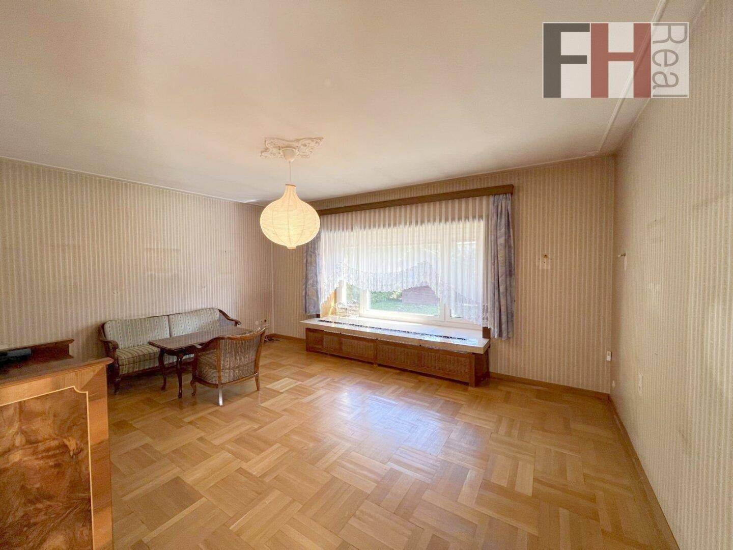 Helles Wohnzimmer mit Fischgrätparkett, großer Fensterfront und klassischer Deckenleuchte.