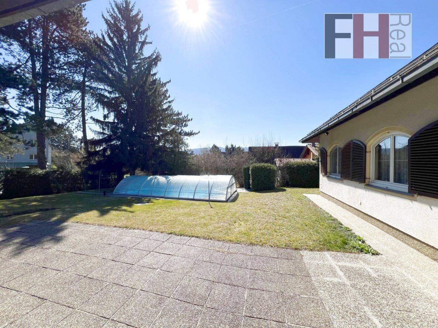 Sonniger Gartenbereich mit gepflasterter Terrasse und überdachtem Swimmingpool.